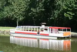 Photo en couleurs. Le bateau-promenade l'Oussance, vu de côté, est amarré sur la berge enherbée du port de Pont-aux-Moines, sur la commune de Mardié. Toute la partie avant est fermée et vitrée, permettant aux touristes de voir le paysage. L’arrière est constitué d’un pont ouvert recouvert d’une bâche rouge. Une large bande rouge tout le long du bateau, blanc par ailleurs, marque la ligne de flottaison.