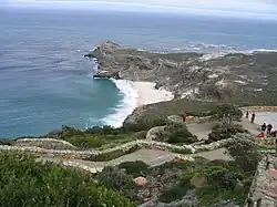 Au premier plan, une allée gravillonnée bordée de murets de pierre serpente sur une pente buissonnante. En fond, un cap rocheux s'avance en biais dans la mer qui s'étend jusqu'à l'horizon brumeux