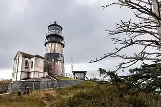 Photo couleur du phare. La peinture blanche cerclée de noire est décrépit et abimée par le temps. Le bâtiment est entourée par un grillage pas très haut, on ne voit pas la mer.