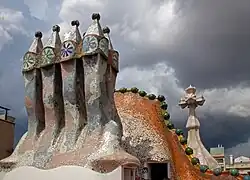 En quatre plans : groupe de cheminées polychromes, toit de la Casa Batlló, croix gaudienne, ciel d'orage.