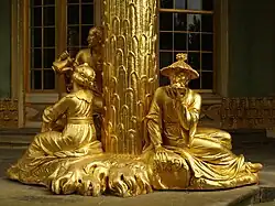 Une sculpture dorée représentant une colonne autour de laquelle évoluent trois personnages, une femme, un homme et une personne portant un chapeau de paille.