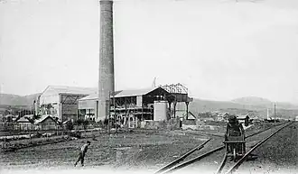 Photo noir et blanc montrant deux bâtiments et une cheminée, une personne pousse une brouette entre des rails.