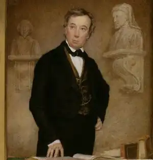 Jeune homme debout, l'air soigné, réfléchi, vêtements marron, légèrement appuyé sur un grand bureau