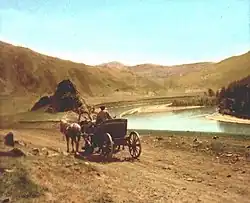 Calèche avec un paysan et deux chevaux longeant une rivière, présumément la Tchouïa, au début du XXe siècle. La scène est représentée sur une carte postale. En arrière-plan se trouve un paysage montagneux.