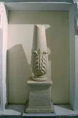Photographie représentant un cippe, petite colonne tronquée en haut, renflée à la base et décorée d'un motif de feuillage. Elle repose sur un socle trapézoïdal dont la face avant porte les inscriptions - en phénicien au-dessus, en grec en dessous.