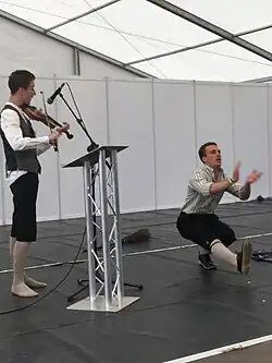 Deux hommes sont sur une scène : l'un joue du violon face à un pupitre, l'autre exécute un numéro de danse.