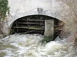 Photographie en couleurs d'une roue en bois sous une arche en maçonnerie.