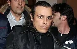 Photographie couleurs d'un homme blanc jeune, cheveux bruns gominés, vêtu d'un blouson de cuir, lors d'une arrestation policière