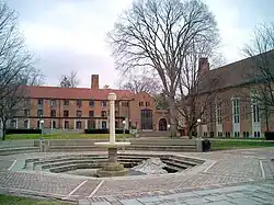 Au premier plan, une fontaine entourée d'un pavement. Au second plan, deux bâtiments perpendiculaires en brique.