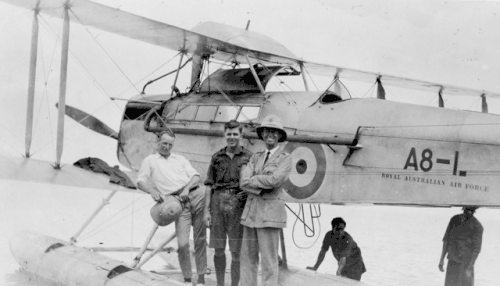Photographie en noir et blanc montrant trois hommes au premier plan. Derrière eux se trouve un avion biplan monomoteur vu de côté avec l'hélice et les ailes à gauche de la photographie. Deux hommes se trouvent derrière l'appareil.