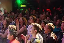 Photographie d'une foule mixte tournée vers la scène. Plusieurs personnes ont des expressions de joie, d'intérêt ou de surprise.