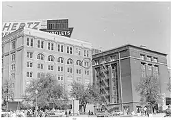 Deux grands bâtiments côte à côte, devant, trois arbres, une foule et des voitures