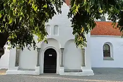 Église blanche sous un arbre, façade simple centrée sur l’entrée à colonnes, dans une lumière douce.