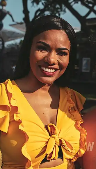 Une femme souriante, au teint mat, cheveux noirs, avec un haut jaune assez décolleté. La personne ferme légèrement les yeux, elle est peut-être face au soleil