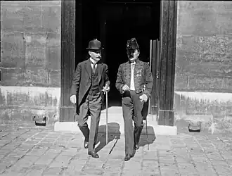 Photo en noir et blanc de deux hommes en train de marcher, épée de l’Académie française à la main : celui de droite a une moustache blanche, un costume trois pièces avec chemise à faux col et chapeau melon ; l’autre, moustache foncée, porte l’habit vert de l’Académie