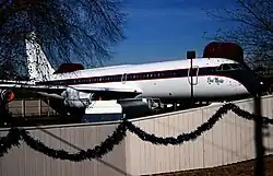 Photo couleur d'un avion aux couleurs blanches avec de fines bandes noires et rouges, entreposé au sein d'un enclos entouré d'un portail blanc.