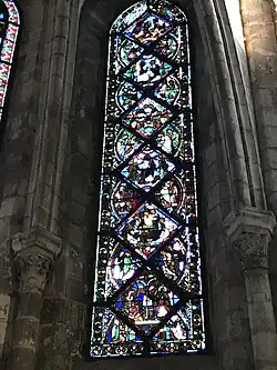 Vitrail composé de pièces rectangulaires et triangulaires. Sont représentés différents personnages dans plusieurs scénettes, difficilement visibles en raison du noircissement du vitrail.