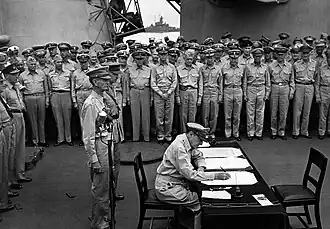 MacArthur est assis et écrit sur un petit bureau. Deux hommes en uniforme se tiennent derrière lui et une large foule d'hommes en uniformes observe la scène.