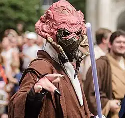 Alien de couleur chair portant un masque respiratoire, des crocs et portant une toge.