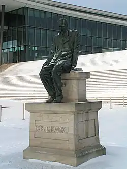 Dostoïevski assis sur le socle statuaire, une main sur son genou droit, tête inclinée vers le sol.