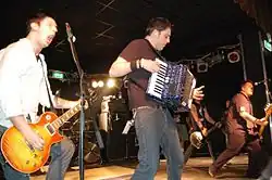 Trois hommes caucasiens au premier plan tenant respectivement, de gauche à droite, une guitare, un accordéon et une guitare. Un autre homme caucasien tenant une guitare au dernier plan est aussi perçu.