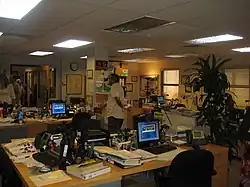 Photo prise à l'intérieur d'un bâtiment présentant une série de bureaux avec des ordinateurs et du papier au sein d'un open-space. Des personnes de l'équipe technique sont présentes à l'arrière-plan.