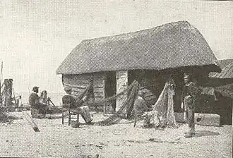 Réparation des filets, sur les galets d'Étretat au pied d'une caloge, bateau de pêche couvert d'un toit, transformé en appentis pour ranger le matériel.