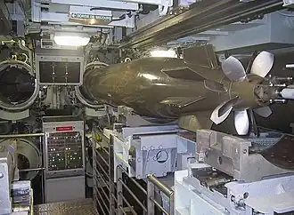 Une pièce encombrée dans laquelle se trouve à droite une torpille et au fond deux tubes ouverts.