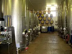 Photographie en couleur montrant des cuves de fermentation en acier inoxydable au Royaume-Uni. Les cuves sont réparties de part et d'autre d'une allée au fond de laquelle on aperçoit des barriques en chêne pour le vieillissement, empilées sur un support contre le mur. Le sol est peint et en pente vers une rigole d'évacuation au milieu du chai.