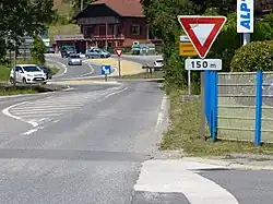 Panneau AB3b avec panonceau M1 «&nbsp;150 m&nbsp;», et panneau AB3a à l'intersection en arrière-plan