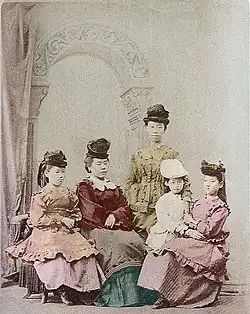 Photo de cinq filles japonaises en costumes de la Belle Époque. Quatre sont assises et une, debout au centre.