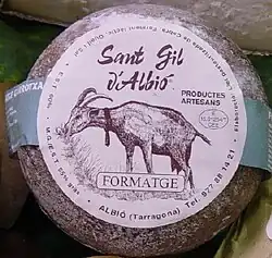 La photographie couleur représente un fromage de chèvre artisanal (indiqué sur l'étiquette) affiné de couleur grise-brun