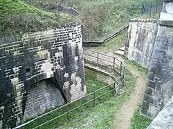 Virage de chemin longeant une barrière, entre deux hauts murs de pierre