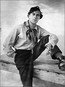 Photo noir et blanc d'un homme en manches de chemise, postérieur et pied gauche sur une table, foulard au cou, cigarette à la main