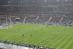 Vue depuis le haut des tribunes d'une rencontre de rugby. L'équipe à droite de l'image s'apprête à faire une remise en jeu, l'équipe opposée étant placée pour la réception.