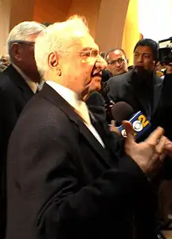 Au premier plan, Frank Gehry de profil est interviewé. Au second plan, caméra et autres journalistes.