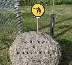 Boule de pierre portant inscription. « Für die Opfer der Unmenschlichkeit ». À l'arrière se trouve une section de clôture et un panneau jaune montrant un soldat agenouillé épaulant son arme.