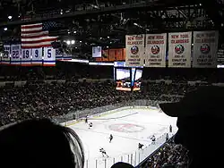 Photo de l'intérieur du Nassau Veterans Memorial Coliseum lors d'un match de hockey dont on aperçoit les joueurs en fond. On voit 4 bannières de champion de la coupe Stanley des Islanders de New York et 5 bannières de maillots retirés de ces mêmes Islanders.