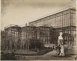 Vue partielle du bâtiment, avec plantations récentes au premier-plan. Un moustachu est appuyé contre le socle d'un buste en pierre. Derrière, une femme en crinoline se promène avec une ombrelle.