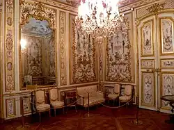 Vue de deux murs d'un cabinet décoré de boiseries blanches et de dorures, couvertes de peintures, avec quatre chaises et un canapé