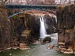 Les gorges d'une rivière, qu'enjambe un pont métallique. Derrière le pont, une chute d'eau.