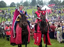 Photographie couleur de deux deux cavaliers parés de pourpre, sur une plaine remplie d'hommes en armure et de spectateurs en habits de ville.