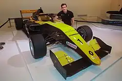 Photographie d'une monoplace de Formule 3 noire et or, vue de trois-quarts dans une exposition, avec un jeune homme à côté.