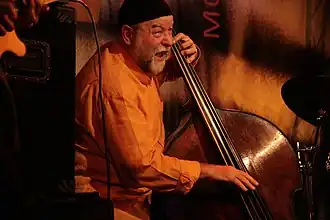un homme âgé, avec une barbe blanche et un petit bonnet, est en train de jouer de la contrebasse sur scène