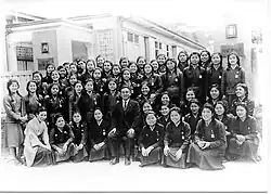 Photo en noir et blanc d'une cinquantaine de jeunes filles asiatiques en uniforme, réparties sur quatre ou cinq rangées ; celles à l'avant sont assises et celles à l'arrière sont debout. Un homme adulte en costume est assis sur une chaise au centre. Un bâtiment scolaire se trouve derrière les filles.