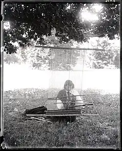 Photo en noir et blanc du travail de We'wha sur un métier à tisser. We'wha est assise dans une prairie. Elle porte un vêtement qui lui arrive aux pieds, dont les manches sont blanches, le reste étant noir. Elle porte un collier similaire à celui qu'elle portait auparavant, mais cette fois-ci certaines petites perles sont pointues. Derrière ou au-dessus de We'wha, on peut voir des feuilles d'arbre et des arbres.