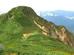 Photo couleur d'un sommet de montagne couvert de verdure, sur fond de ciel blanc laiteux.