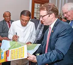 Un homme de type indien, vêtu de blanc et tenant un feutre commente une carte du Sri Lanka que lui tend un élu américain en costume bleu.