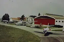Photographie ancienne représentant quatre avions devant un hangar à la porte rouge. De gauche à droite :  un B-17, un Hurel, un AirCommander et un Mystère 20.