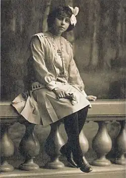 Photographie sépia d'une jeune fille en pied assise sur le rebord d'une balustrade.